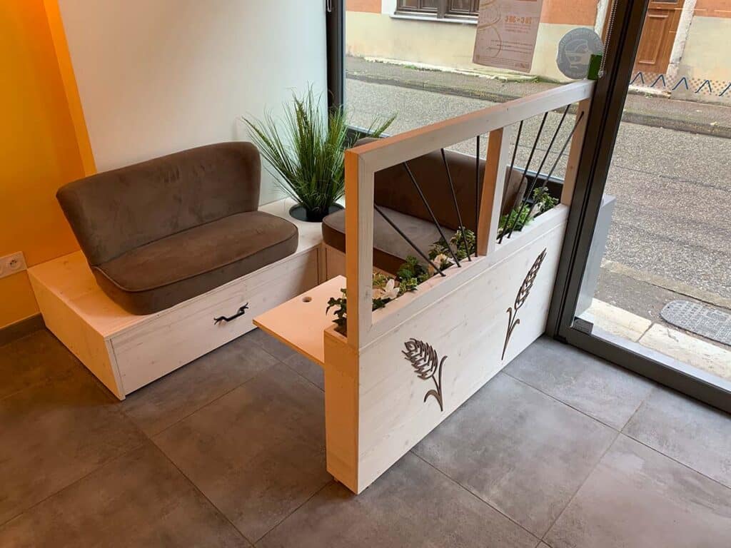 Zone de sièges style boulangerie avec canapé, banquette bois tiroirs, jardinière déco épi blé près fenêtre. Affiche 3J visible.